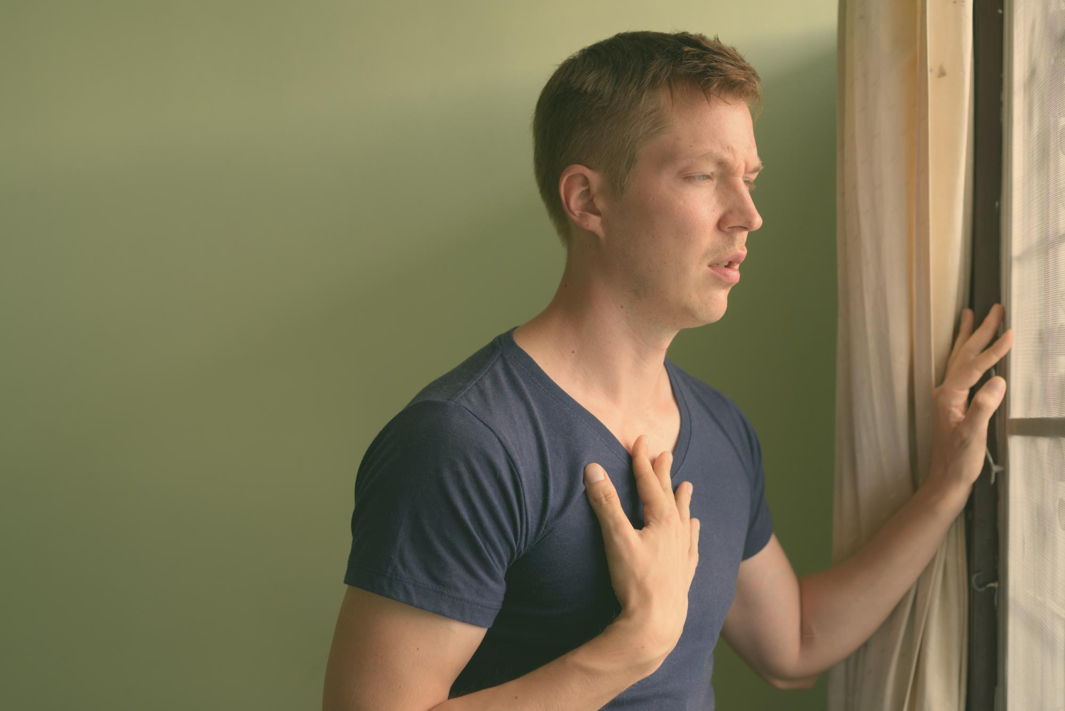 Een man met een blauw t-shirt aan, staat voor het raam en kijkt naar buiten. Hij heeft zijn ene hand tegen het raam aan en zijn andere hand heeft hij op zijn borst. De man oefent met zang- en ademhalingstechnieken.